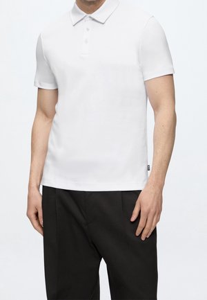 Homme portant un polo blanc uni à manches courtes et un pantalon noir, debout devant un fond blanc.