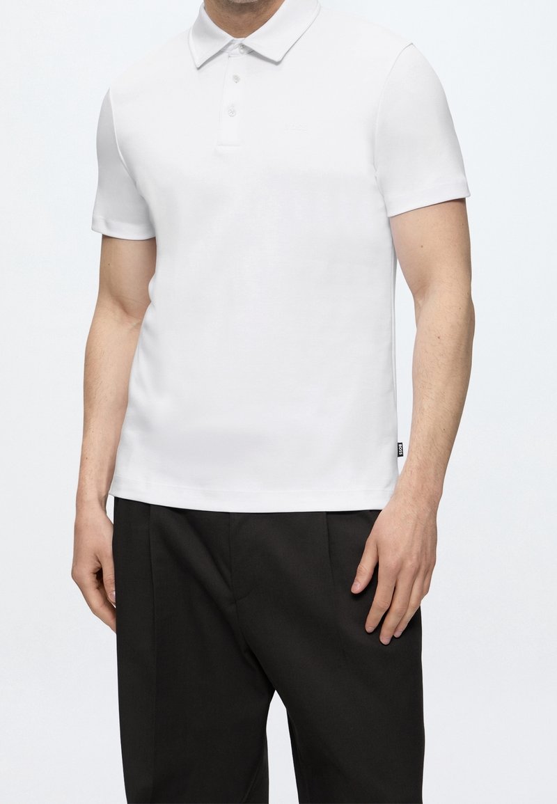 Homme portant un polo blanc uni à manches courtes et un pantalon noir, debout devant un fond blanc.
