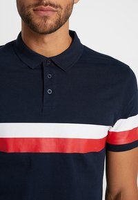 Polo azul marino de algodón con cuello, con rayas horizontales en blanco y rojo. Cinco botones y una textura suave.
