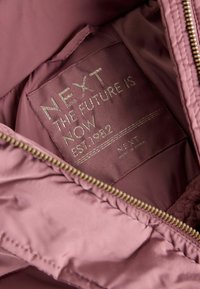 Etiqueta dentro de una chaqueta malva con texto dorado que dice "NEXT THE FUTURE IS NOW EST. 1982" y una cremallera dorada parcialmente visible.