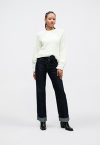 Bijeli teksturirani džemper s dugim rukavima, uparen s tamnim trapericama širokih nogavica ukrašenim remenom u obliku ruže. Crne gležnjače zaokružuju izgled.