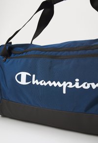 Marineblå duffelbag laget av teksturert stoff, med svart bunn, topplukkeren og iøynefallende hvit "Champion"-logo på siden.