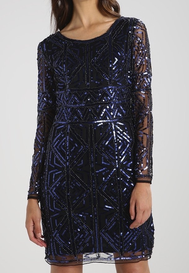 Femme portant une robe noire ajustée avec des manches longues transparentes décorées de motifs géométriques en sequins bleus et noirs.