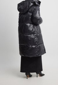 Schwarzer Puffer-Mantel mit glänzender Oberfläche, Kapuze und steppdesign. Hat einen langen Schnitt und wird mit einem schwarzen Jeansrock und spitzen High Heels kombiniert.