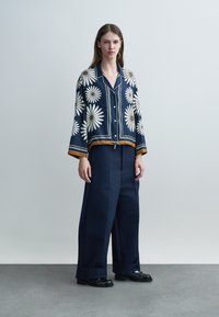 Γυναίκα που φοράει μπλε πουκάμισο με κουμπιά και σχέδιο από μαργαρίτες, με μουσταρδί λεπτομέρειες, φαρδιά μπλε παντελόνια και μαύρα παπούτσια, στέκεται μπροστά σε απλό φωτεινό φόντο.