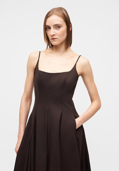 Jovem mulher com cabelo castanho claro até aos ombros, vestindo um vestido castanho escuro sem mangas com alças finas, em pé com uma mão dentro do bolso.