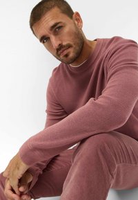 Pull en tricot rose avec texture côtelée, associé à un pantalon en velours côtelé assorti. Modèle assis, les bras croisés, sur un fond neutre.