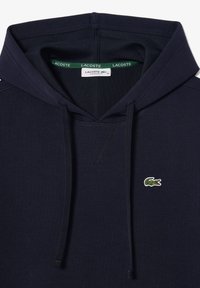 Tamsiai mėlyna džemperis su tekstūruota medžiaga, turintis su varpsteliu reguliuojamą gobtuvą ir mažą žalią bei baltą Lacoste krokodilo logotipą krūtinėje.