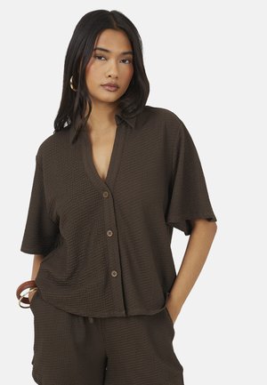 Femme aux longs cheveux foncés portant une chemise texturée marron foncé à manches courtes boutonnée et un short assorti, mains dans les poches, expression neutre.