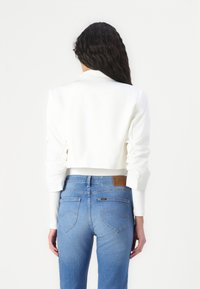 Krojované bílé sako s hladkou texturou a řasenými rukávy, spojené s modrými džíny se středním pasem a koženou značkovou nášivkou na zadní straně.