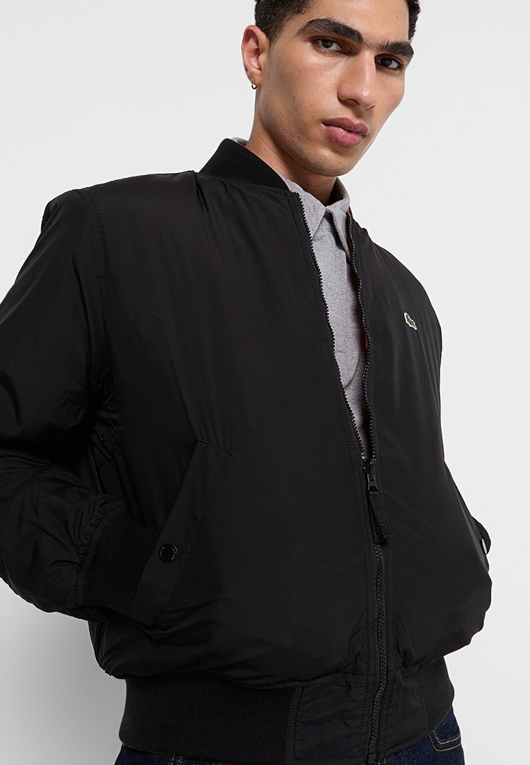 Veste bomber noire avec fermeture éclair sur le devant, poignets et col côtelés. Présente un petit logo sur la poitrine et une texture douce et lisse.