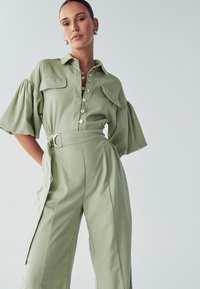 Moteris vilki šalavijo žalią kombinezoną su diržu, pūstomis trumpomis rankovėmis ir sagomis priekyje, stovinti prieš paprastą šviesų foną.