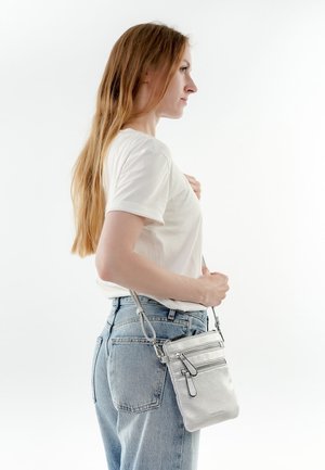 Bolso cruzado de cuero plateado con superficie texturizada, dos cremalleras frontales y una correa ajustable, llevado por una persona vestida con una camiseta blanca y jeans.