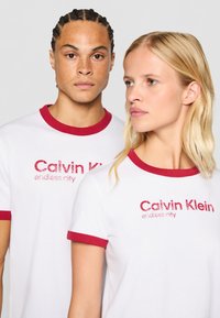 Balti marškiniai su raudonais ribotais apykaklėmis, su užrašu „Calvin Klein endless city“ nusidėvėjusiais raudonais rašmenimis. Lygus audinys su prigludusia forma.