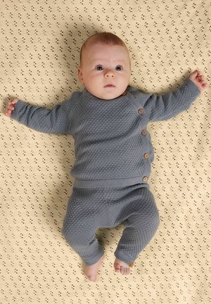 Baby liggend op een crèmekleurige gebreide deken, gekleed in een grijze gebreide outfit met houten knopen, armen gespreid en omhoog kijkend.
