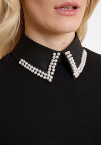 Haut en tissu noir avec un col structuré orné d'accents en perles blanches disposés en forme de V le long de la ligne du col.