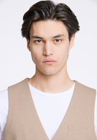 Ljusbeige väst över en vit skjorta, med en subtil textur och en enda bröstficka, i en klassisk skärning och rena linjer.