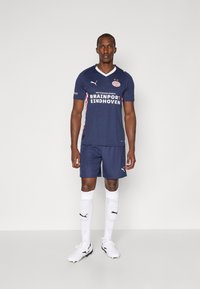 Blå fotbollsdräkt med vita detaljer och "PSV EINDHOVEN"-logotyp, ihop med blå shorts och vita strumpor med svart Puma-logga.