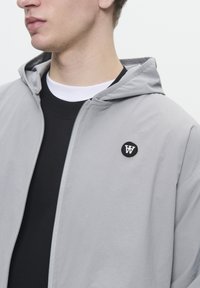 Lys grå hættejakke med lynlås og sort broderet logo på brystet. Båret over en sort skjorte med hvid krave.