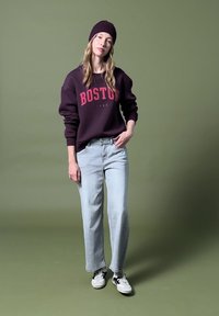 Ung kvinde iført en mørk lilla Boston-sweatshirt, matchende hue, lyseblå jeans og hvide sneakers, stående mod en olivengrøn baggrund.