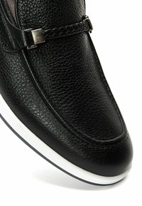 Mocassin en cuir noir avec une finition texturée, doté d'une lanière tressée et de ferrures argentées. Semelle blanche avec un léger accent bleu.