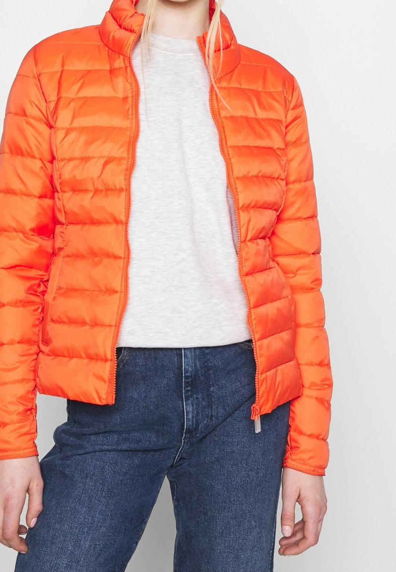 Lys orange dunjakke med vandret quiltning, høj krave og lynlås, kombineret med en lysegrå skjorte og mørke denimjeans.