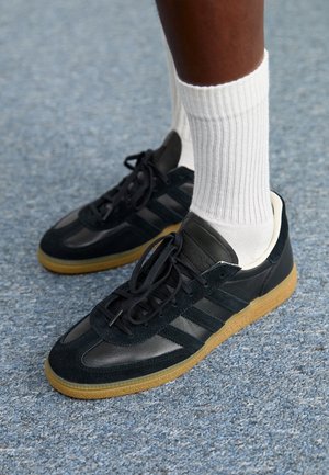 Persoon die zwarte leren en suède sneakers met gumzolen en witte geribde kousen tot aan de kuit draagt, staand op een blauw tapijt.