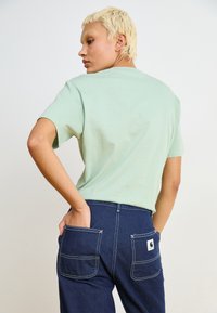 Mintgrön kortärmad t-shirt i avslappnad passform, kombinerad med mörkblå byxor med två stora bakfickor och kontrasterande vit sömnad.