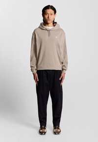 Beige hoodie med dragsko, med en liten logotyp på bröstet. Parat med svarta lösa byxor och mönstrade sneakers.