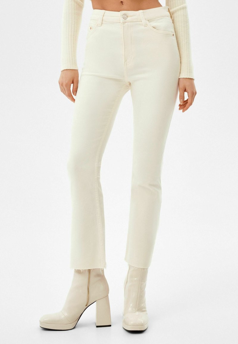 Jean blanc taille haute à coupe slim, avec des détails d'ourlet effiloché et cinq poches, associé à des bottines à talons brillantes.