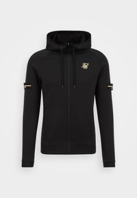 Sudadera con cremallera negra hecha de tela suave, con un logo dorado en el pecho, bolsillos con cremallera y detalles decorativos en las mangas.