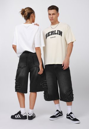 Svarta denimshorts med fransade kanter och slitna detaljer, i kombination med en vit t-shirt och en krämfärgad grafisk t-shirt, tillsammans med sportiga skor.