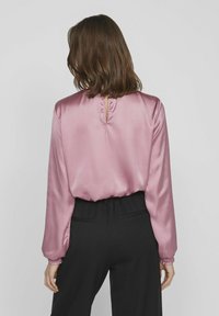 Långärmad satinblus i mjuk rosa, med avslappnad passform, volangärmar och en liten nyckelhålsspringa i bakhalsen. Matchad med svarta byxor.