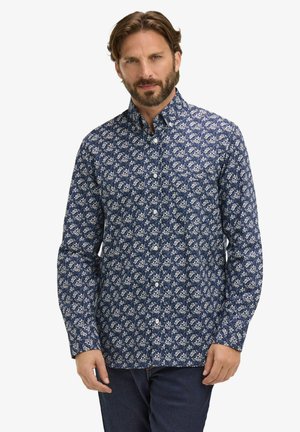 Homme portant une chemise à manches longues bleue à boutons avec un petit motif floral blanc et un jean bleu foncé, debout devant un fond blanc.