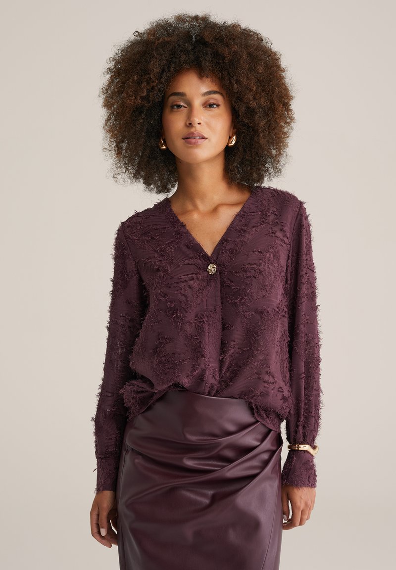 Violettpälsad blus med långa ärmar, V-ringning och knapprad, tillsammans med en figurnära, mörk lila lässkirt.
