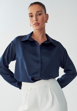 Navy blue satin button-up shirt with a classic collar, long sleeves, and a relaxed fit, paired with white high-waisted trousers.