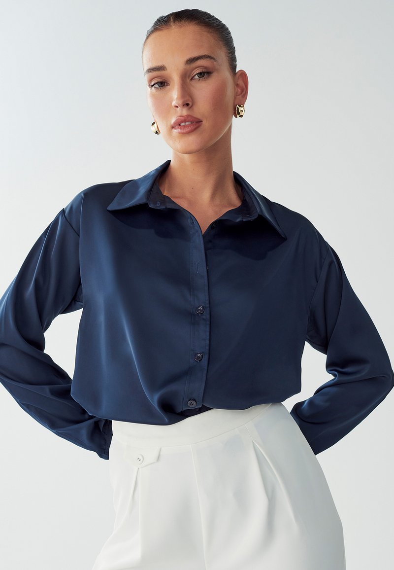 Camicia in satin blu navy con bottoni, colletto classico, maniche lunghe e vestibilità rilassata, abbinata a pantaloni bianchi a vita alta.