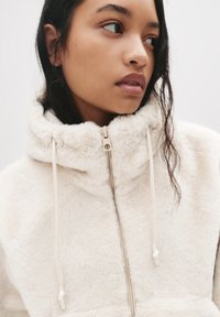 Svetlo bež flis jakna z visokim ovratnikom, sprednjim zadrgo in vrvicami. Mehka tekstura z minimalističnim dizajnom in brez vidnih vzorcev.