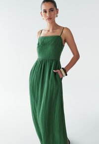 Zaļš maksi kleita ar strukturētu augšdaļu, plānām siksnām un teksturētu audumu. Iekļauj saspringtu jostasvietu un sānu kabatas, kas nodrošina funkcionalitāti.