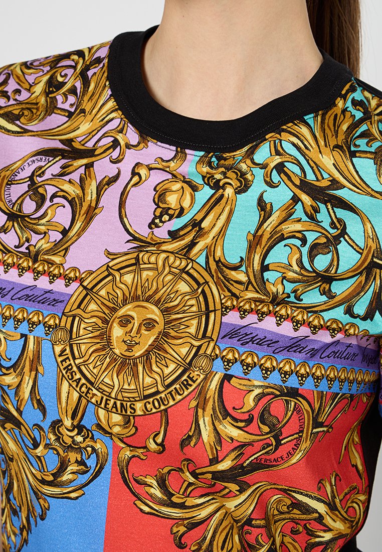 Koszula krótki rękaw w wielokolorowe wzory z ozdobnymi złotymi motywami, czarnym dekoltem i emblematem słońca. Wykonana z miękkiego materiału o gładkiej teksturze.