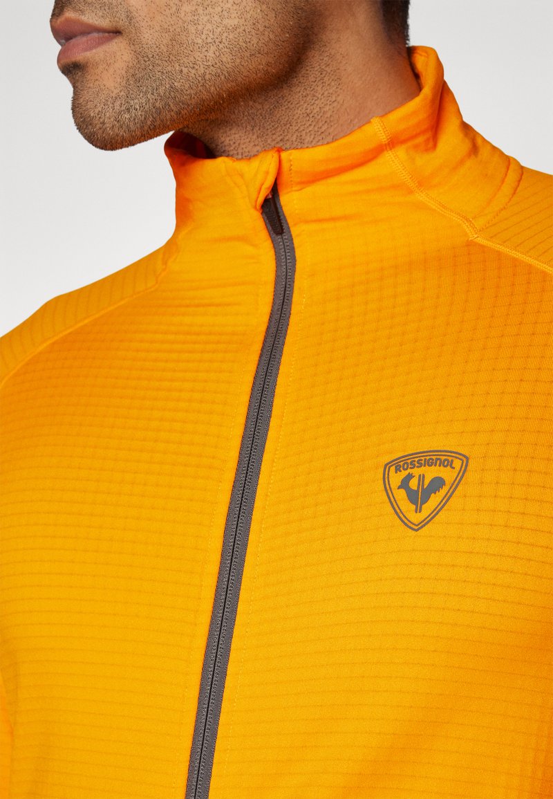 Oranje zip-up jack van textiel met een grijze rits en een logo patch. Hoge kraag ontwerp.