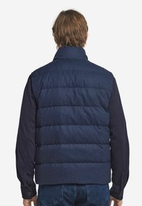 Marineblaue Steppweste mit hohem Kragen, ausgestattet mit horizontalen, gesteppte Paneelen und einer glatten Stofftextur, kombiniert mit dunklen langen Ärmeln.