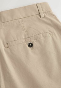 Pantalon en tissu beige avec une poche passepoilée à l’arrière fermée par un bouton noir et un passant de ceinture à la taille.
