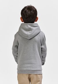 Grå hættetrøje med snørelukning på hætten, lavet af et blødt, tekstureret stof. Synlige syninger og en løs pasform; båret over beige bukser.