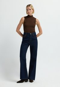 Top sin mangas de punto marrón con costuras acanaladas, combinado con pantalones de mezclilla oscuros de tiro alto y pernera acampanada. Notables costuras de contraste y detalles de bolsillo en marrón.