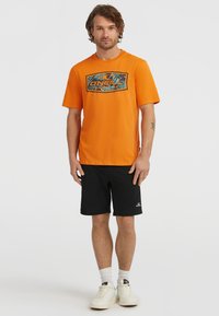Orange cotton t-shirt with a graphic print, paired with black shorts and white sneakers. The shirt features a crew neckline and short sleeves.