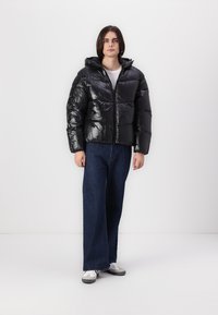 Sort, skinnende pufferjakke med hætte, udstyret med lynlås og quiltet design. Båret med blå vide jeans og hvide sneakers.