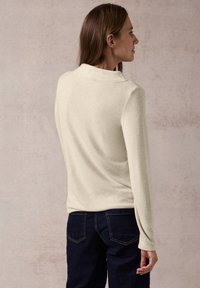 Helles beige Langarm-Sweatshirt mit strukturiertem Finish, hochgeschlossen und figurbetont, kombiniert mit dunklen Jeans.
