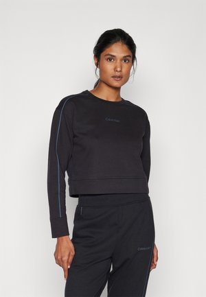 Musta lyhytnakki sweatshirt, jossa on sininen korostustikkaus, pyöreä pääntie ja Calvin Klein -logo. Pehmeä kangas ja pitkät hihat.