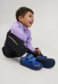 Młody chłopiec śmieje się, siedząc na podłodze, ubrany w jasnofioletową kurtkę, czarne spodnie i niebieskie trampki na rzepy.